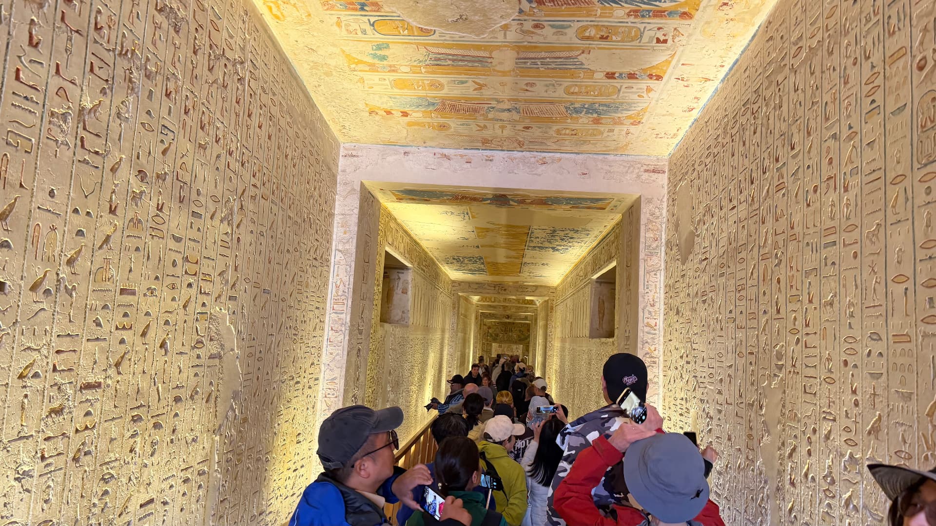 A tomb corridor in the Valley of the Kings, walls covered in hieroglyphs and a painted ceiling, visitors filing through