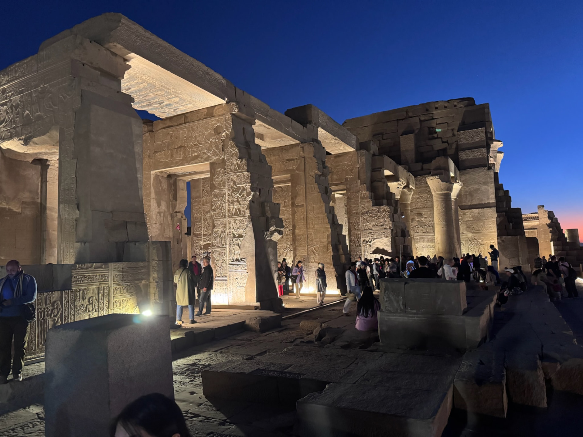 Carved columns and reliefs of Kom Ombo lit from below at dusk with visitors walking between them