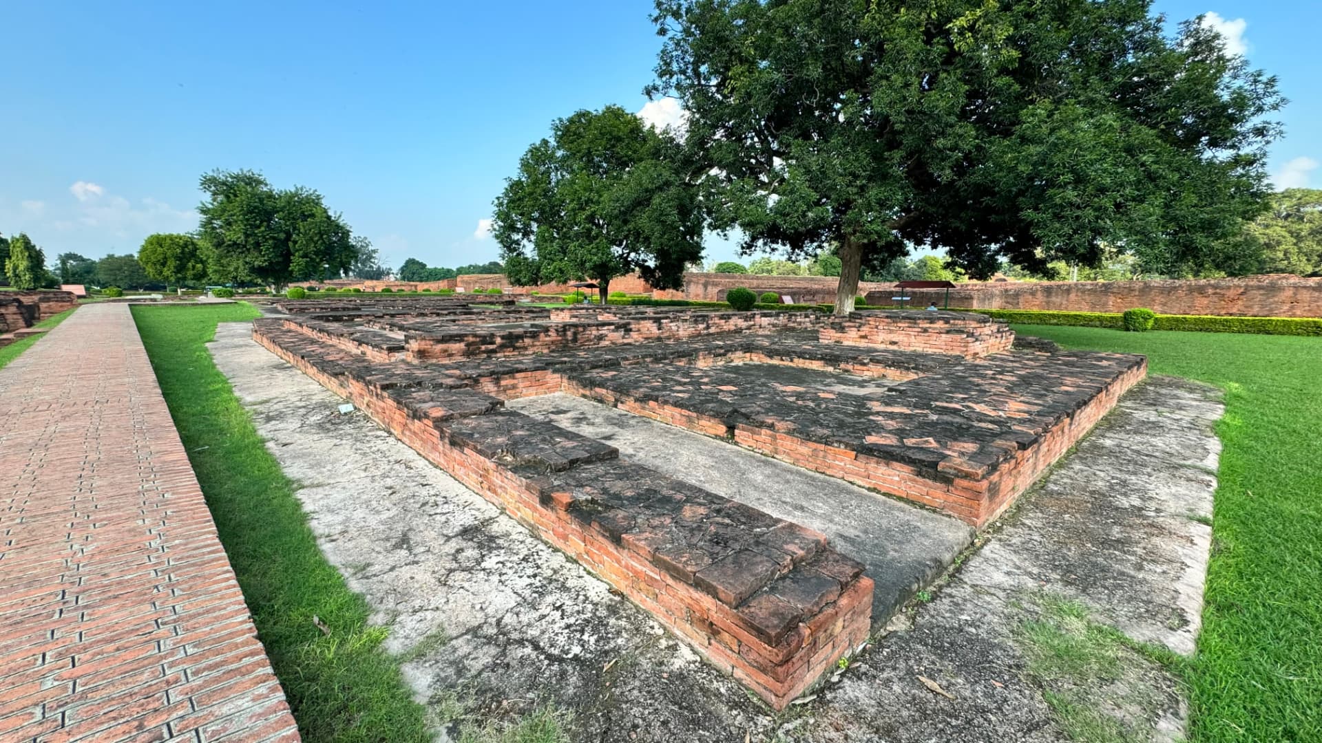 Rectangular brick foundations lie beside a shaded walkway. — Nalanda, Bihar, India · Patna chapter.