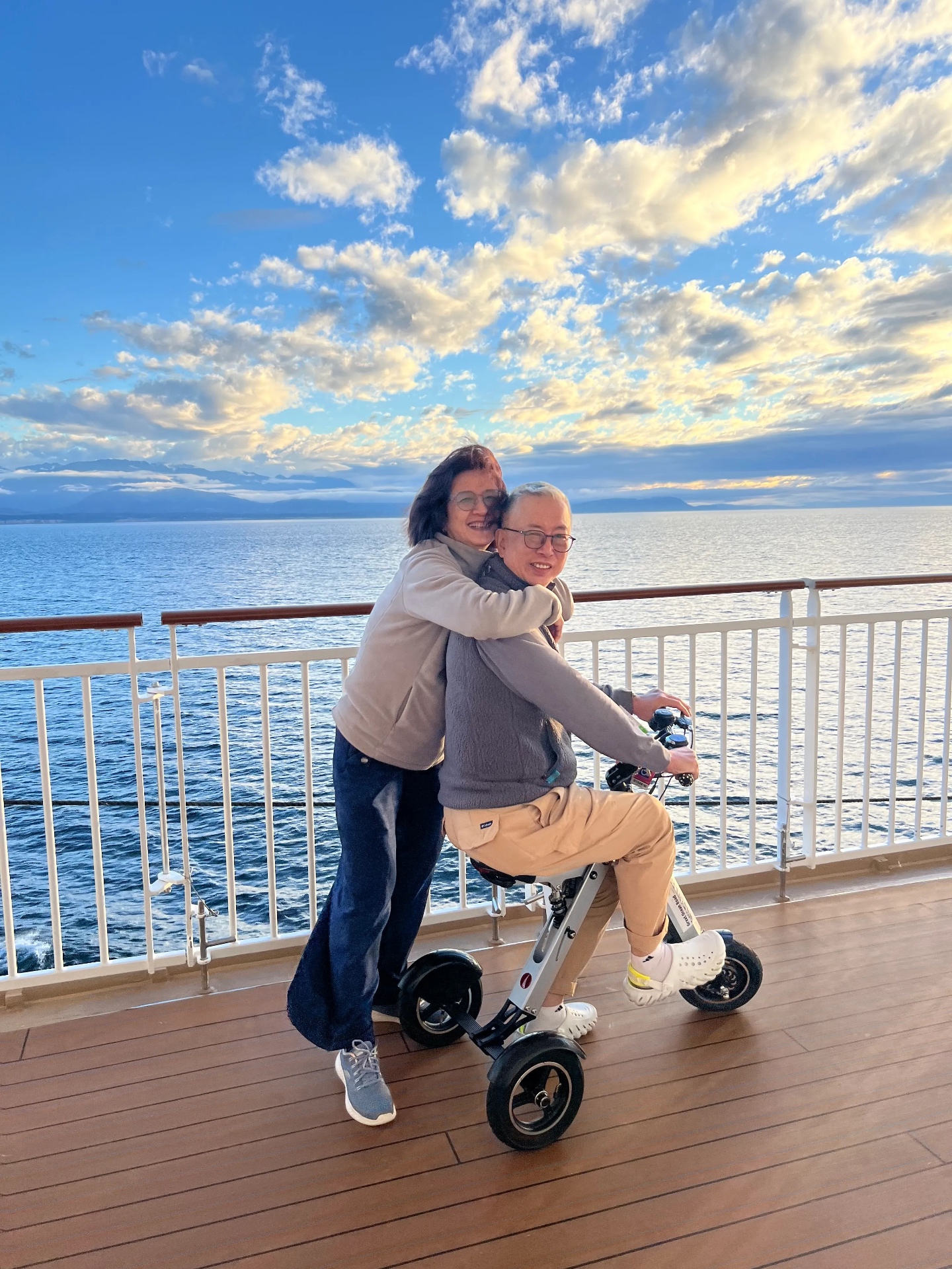 Ken riding his white three-wheeled mobility tri-cycle on the ship's deck at sunset, with Becky standing behind him with her arms around his shoulders, calm sea and mountains behind
