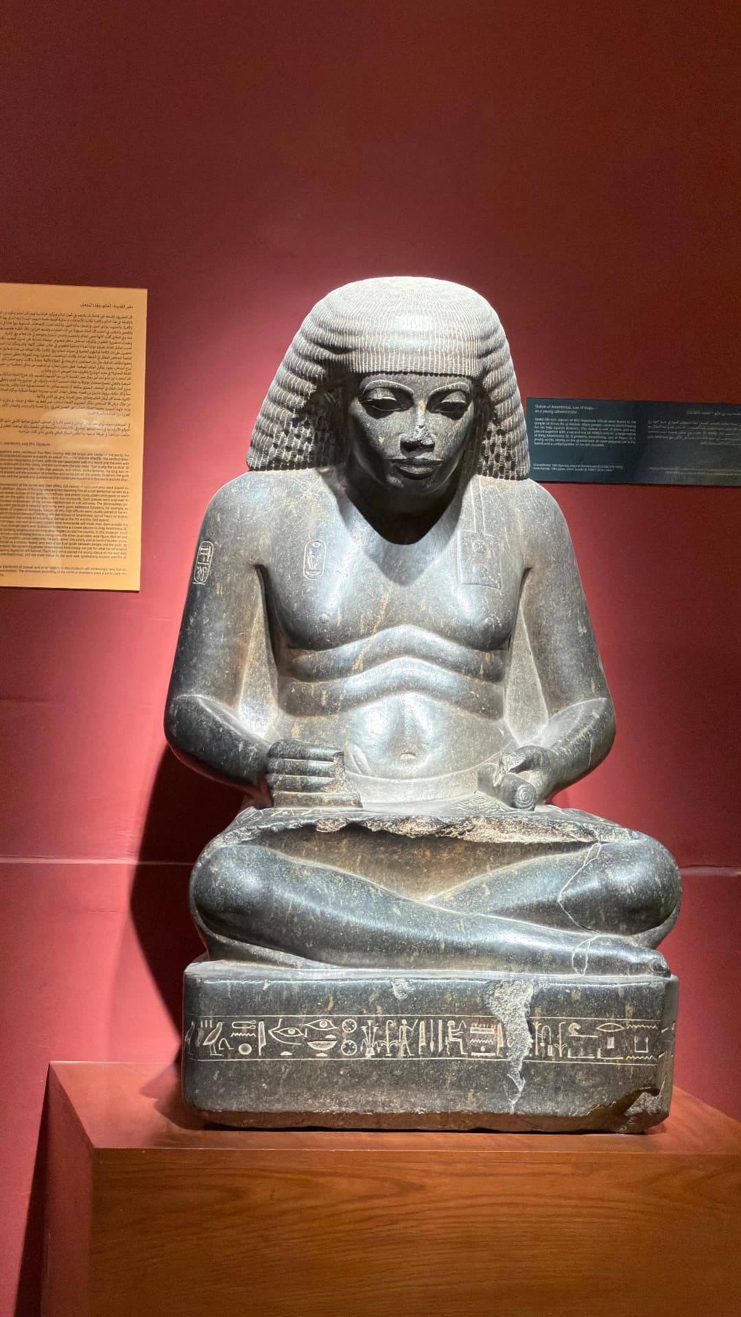 A seated granite statue of a scribe with a scroll across his lap, lit against a red wall