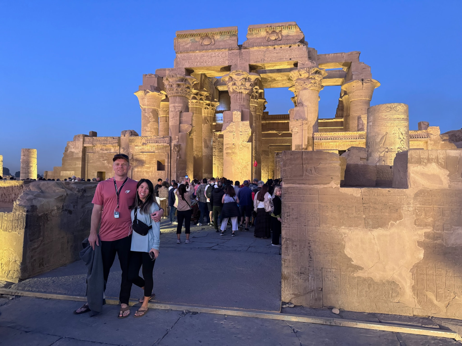 A couple posing at the entrance to the floodlit Temple of Kom Ombo under a deep blue evening sky