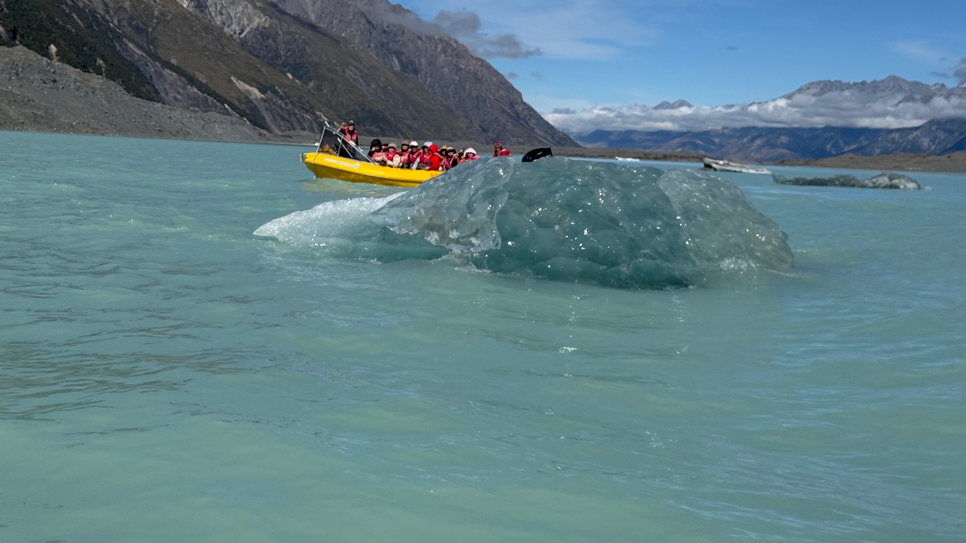 A yellow tour boat full of passengers in red life jackets beside a low turquoise iceberg on the milky lake