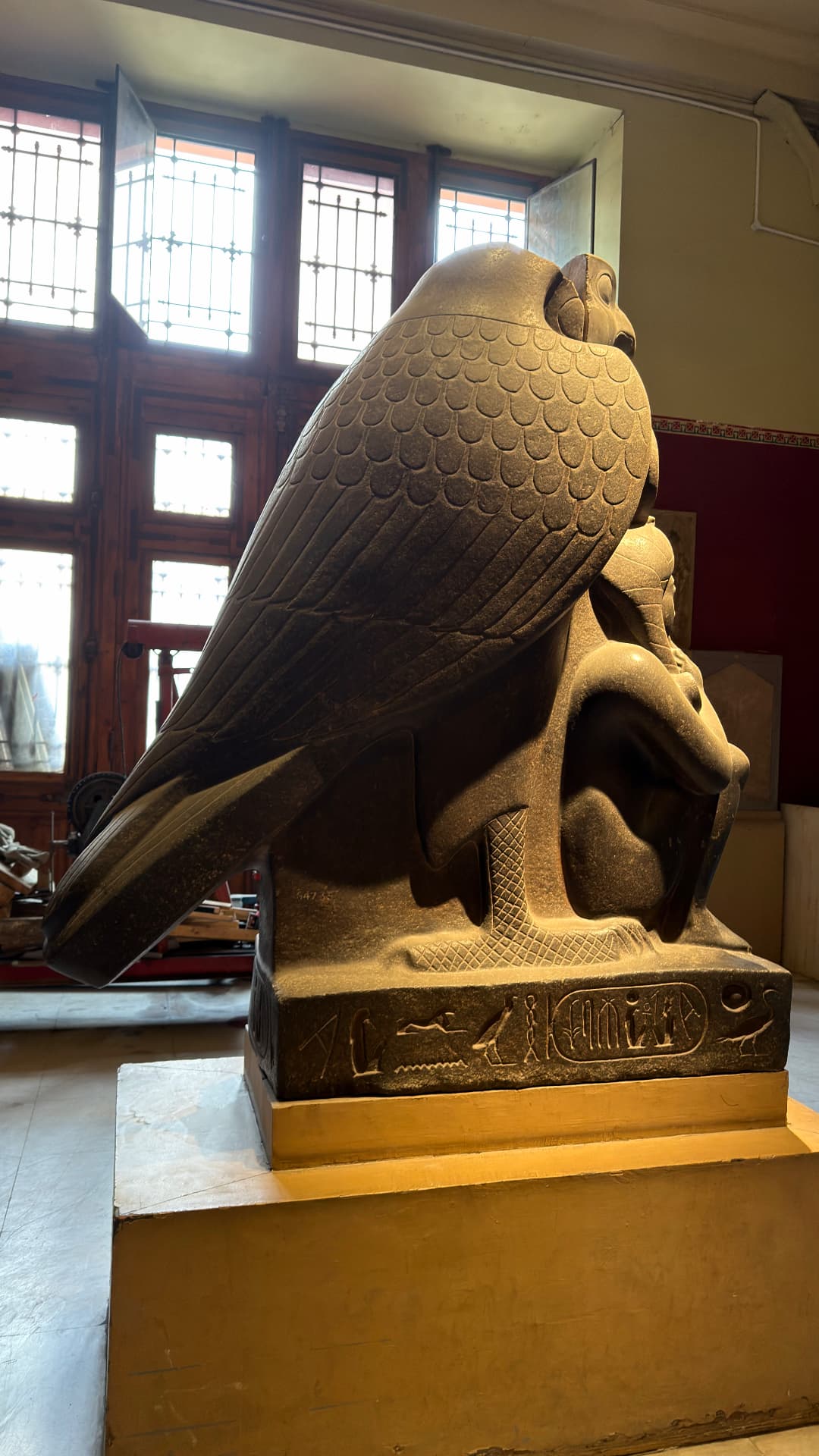 A large granite falcon sculpture sheltering a small pharaoh figure, backlit by tall windows