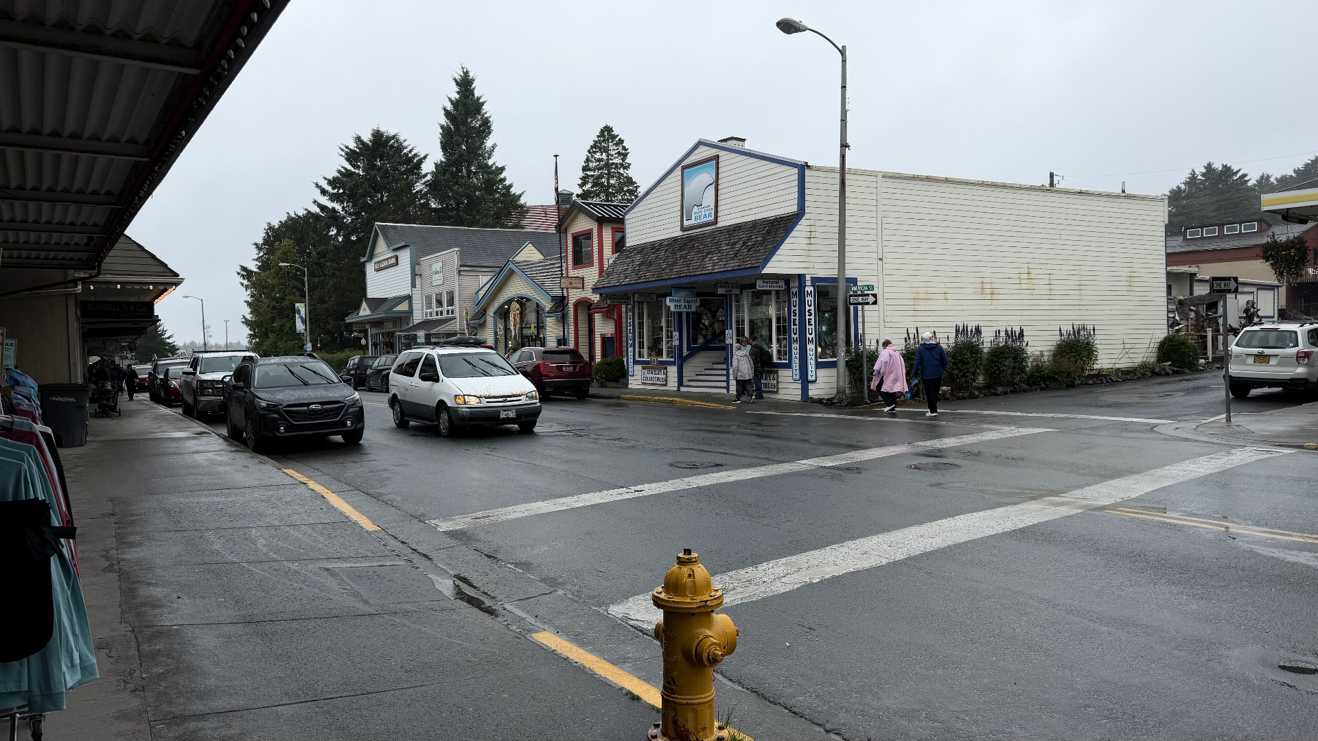 A wet main street in Sitka lined with small shops and galleries, cars parked along the kerb under a grey sky