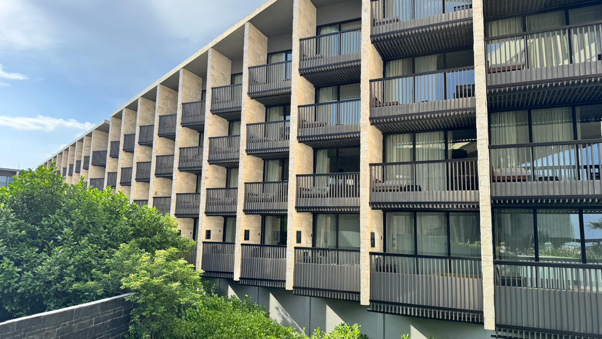 Long hotel facade with shaded balconies above tropical greenery
