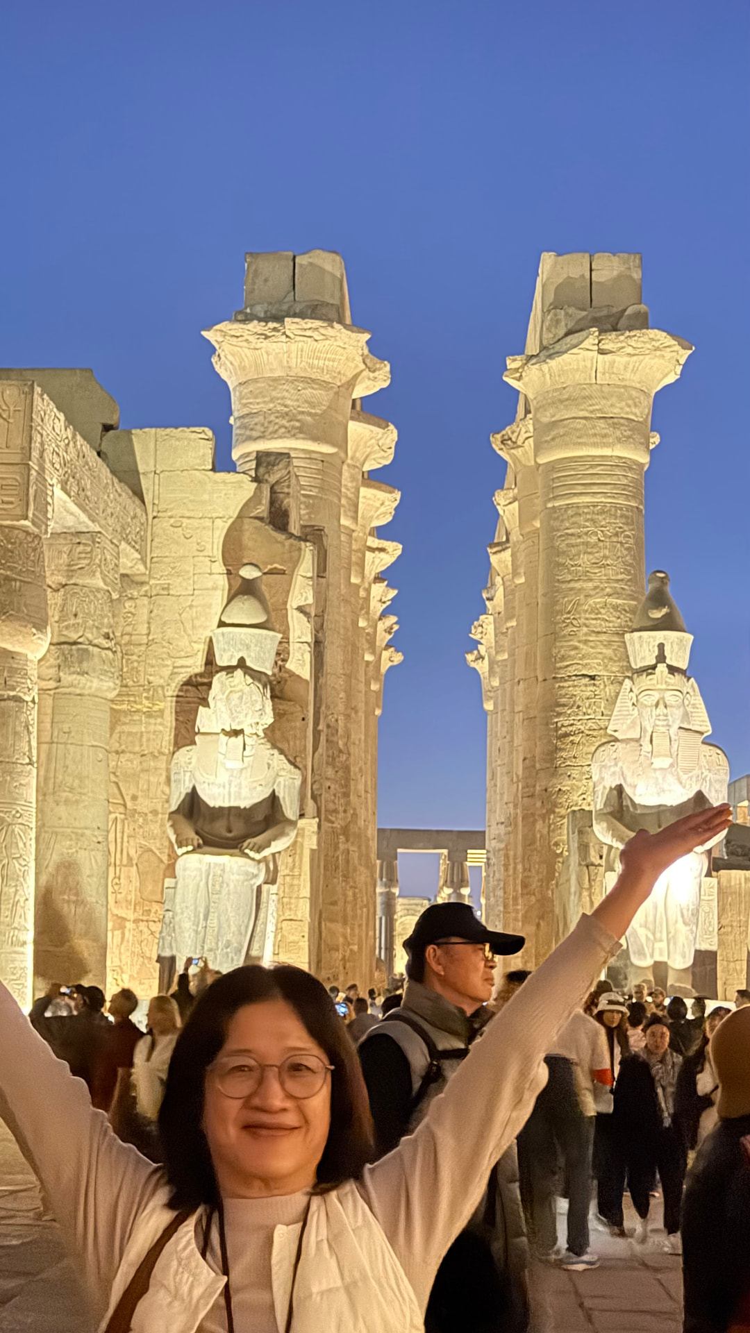 Becky with arms raised in front of the lit colossi and columns of Luxor Temple
