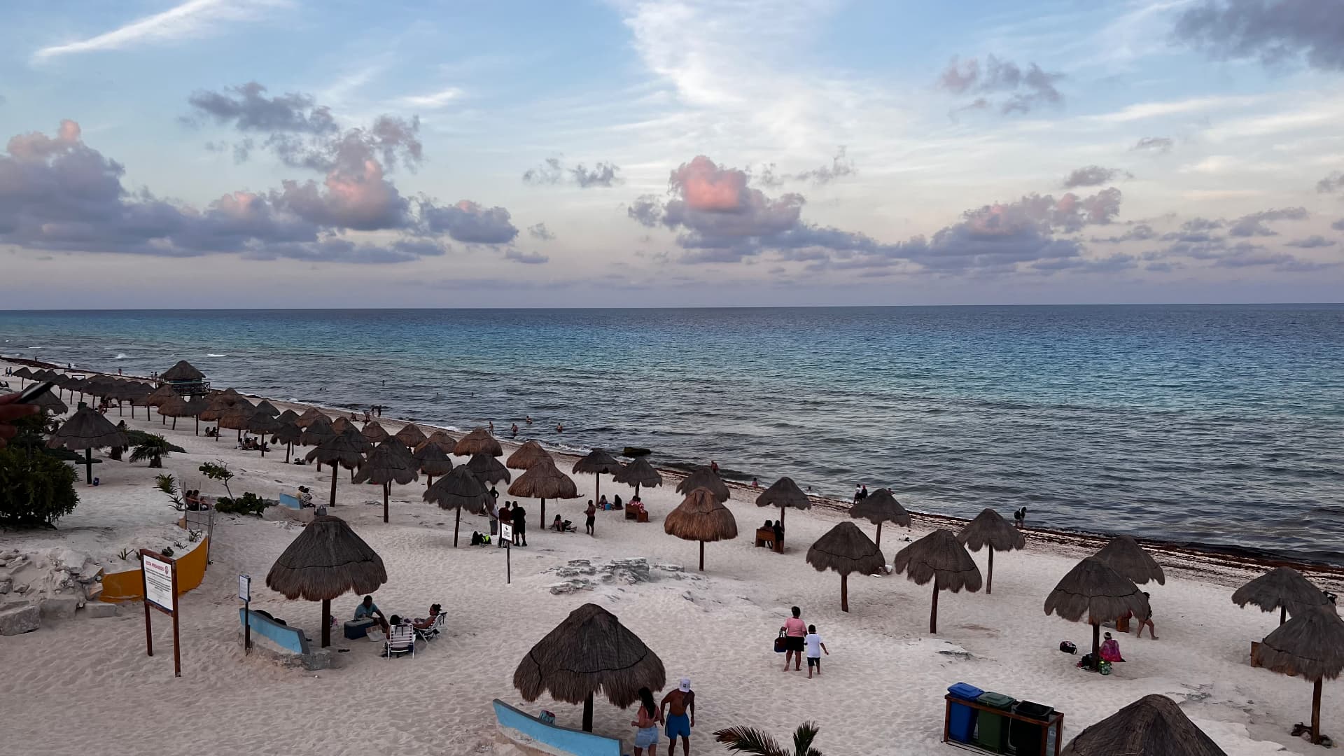 Palapa umbrellas along a broad beach beneath pastel clouds