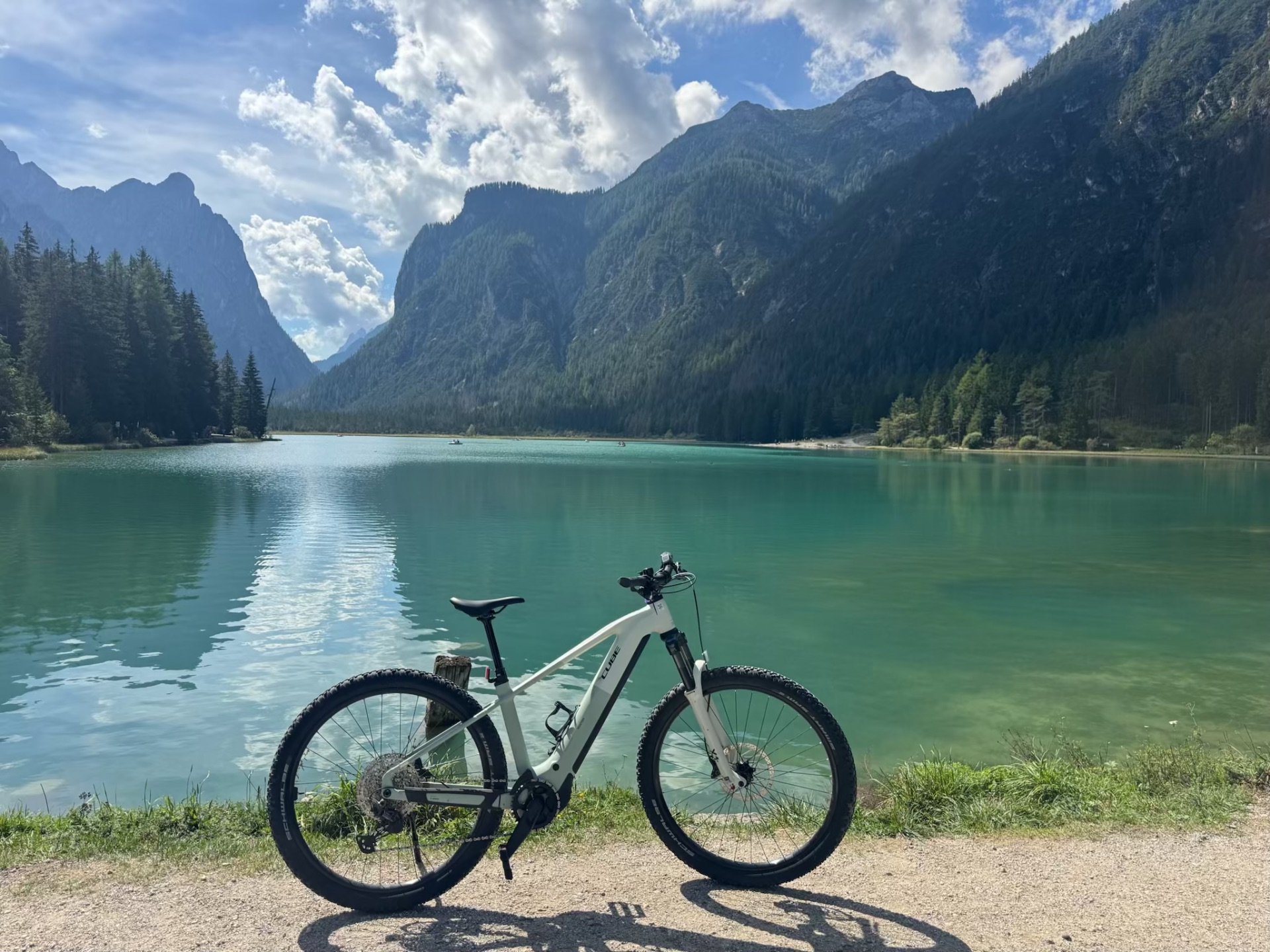 A white mountain bicycle beside a turquoise alpine lake backed by forested peaks