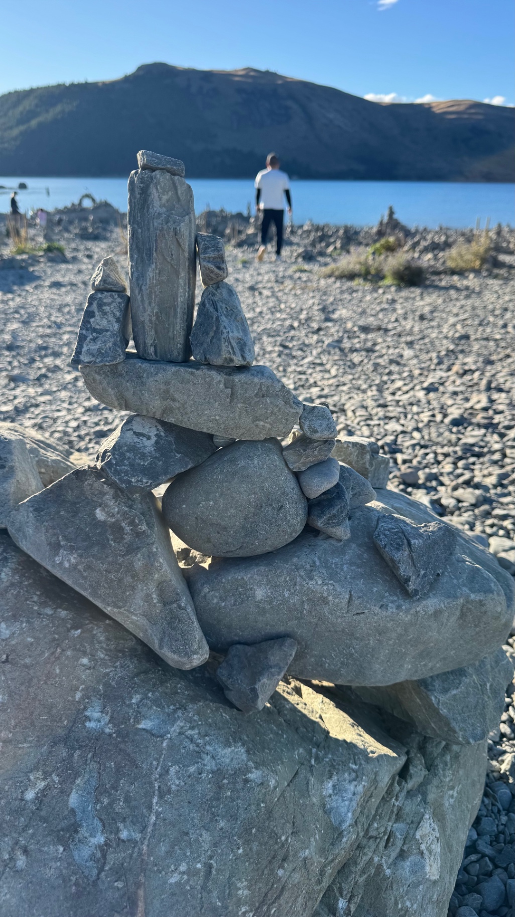A close-up of a carefully balanced stone cairn on a boulder, with the lake and hills behind