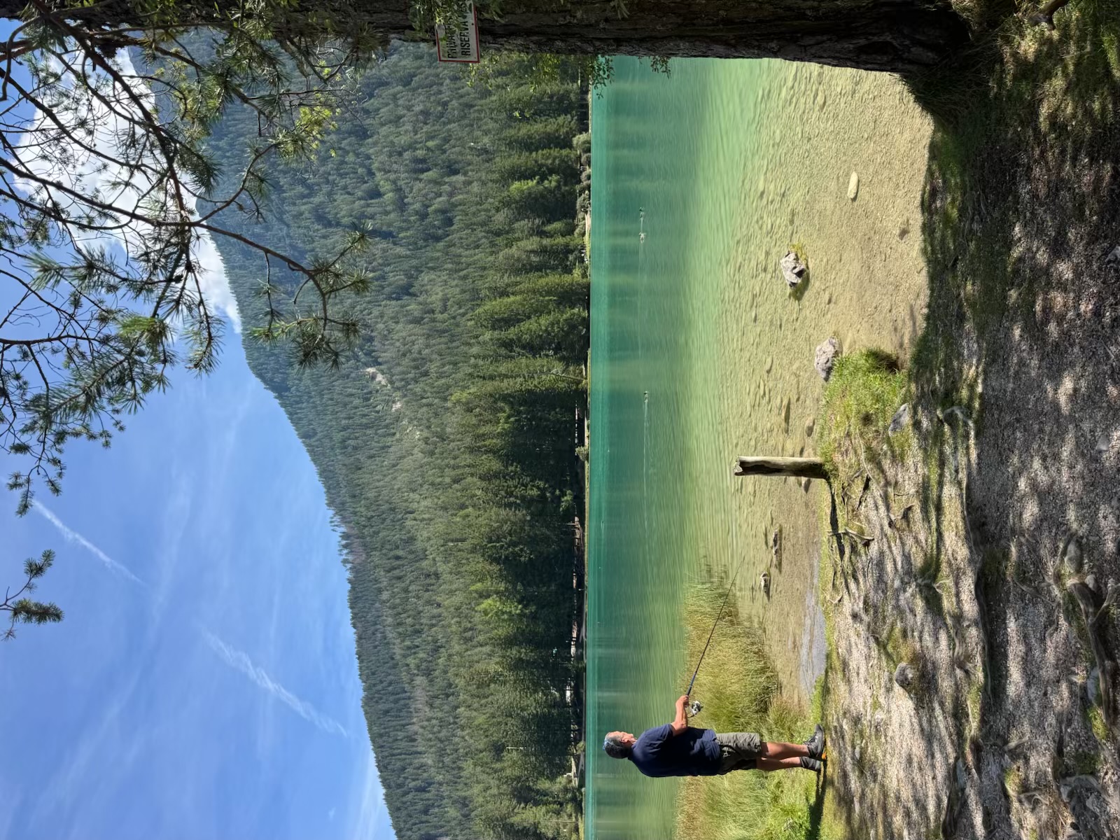 Ken fishing from the wooded shore of a clear turquoise alpine lake