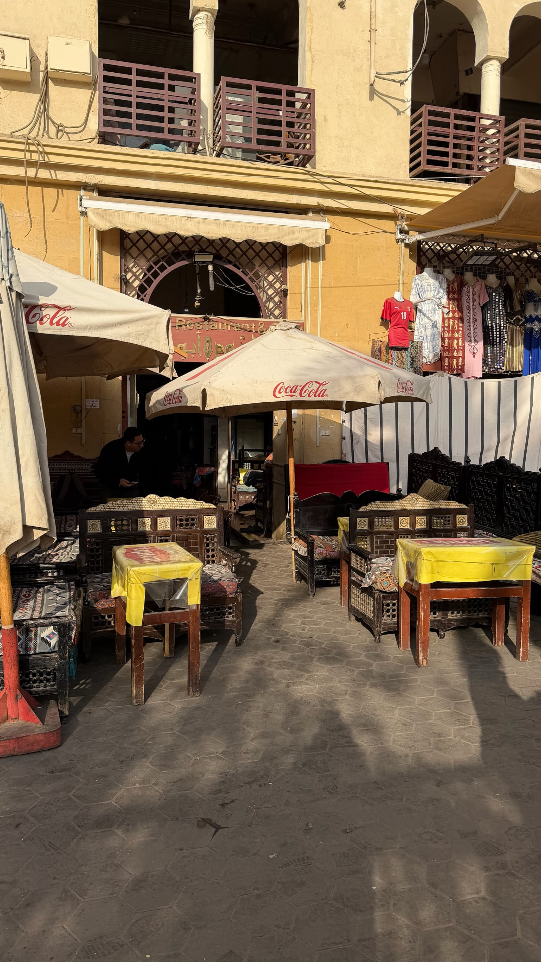 An outdoor café with Coca-Cola umbrellas, carved wooden benches and yellow tables in a sunlit Cairo lane