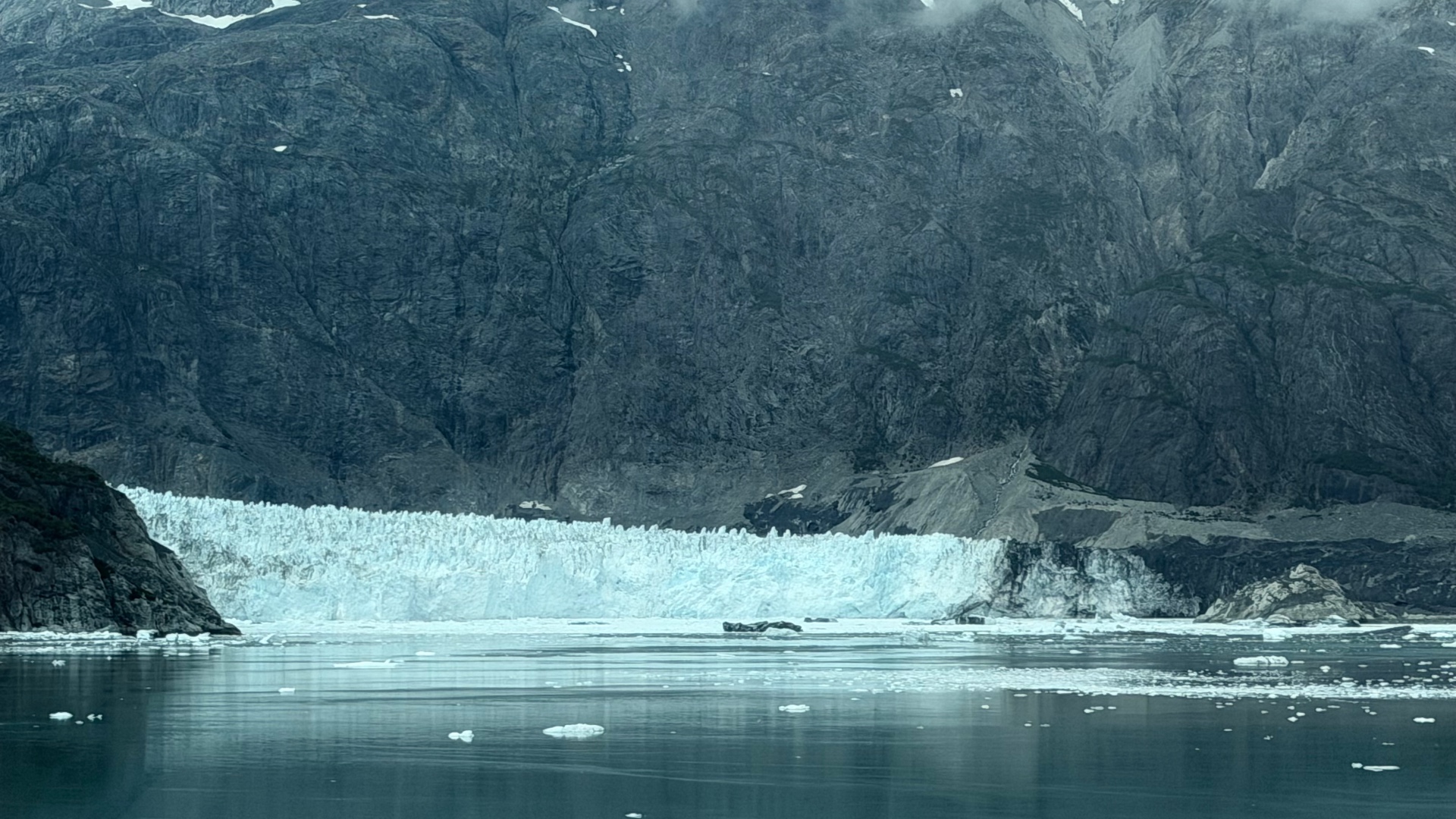 A wide blue glacier fronting dark mountain cliffs across still water