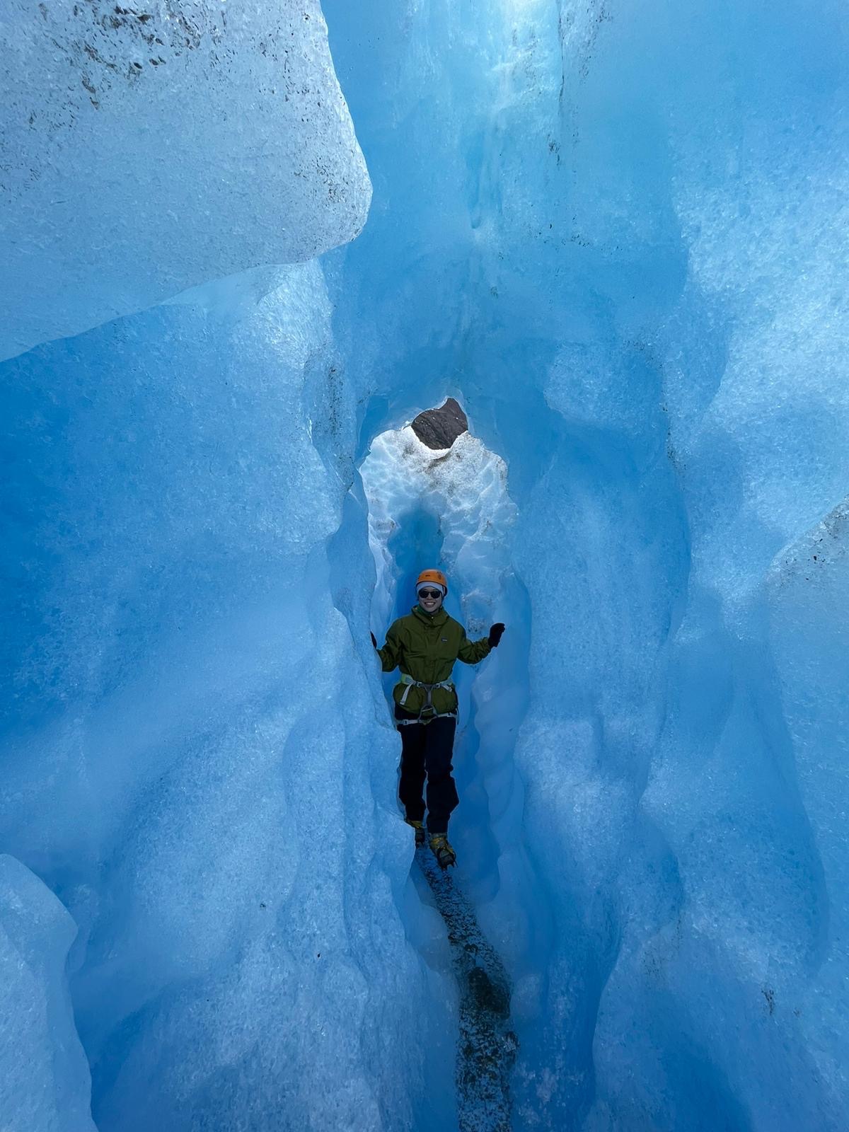 A woman in an olive jacket and orange helmet stands inside a deep narrow ice crevasse, surrounded by glowing translucent blue ice walls