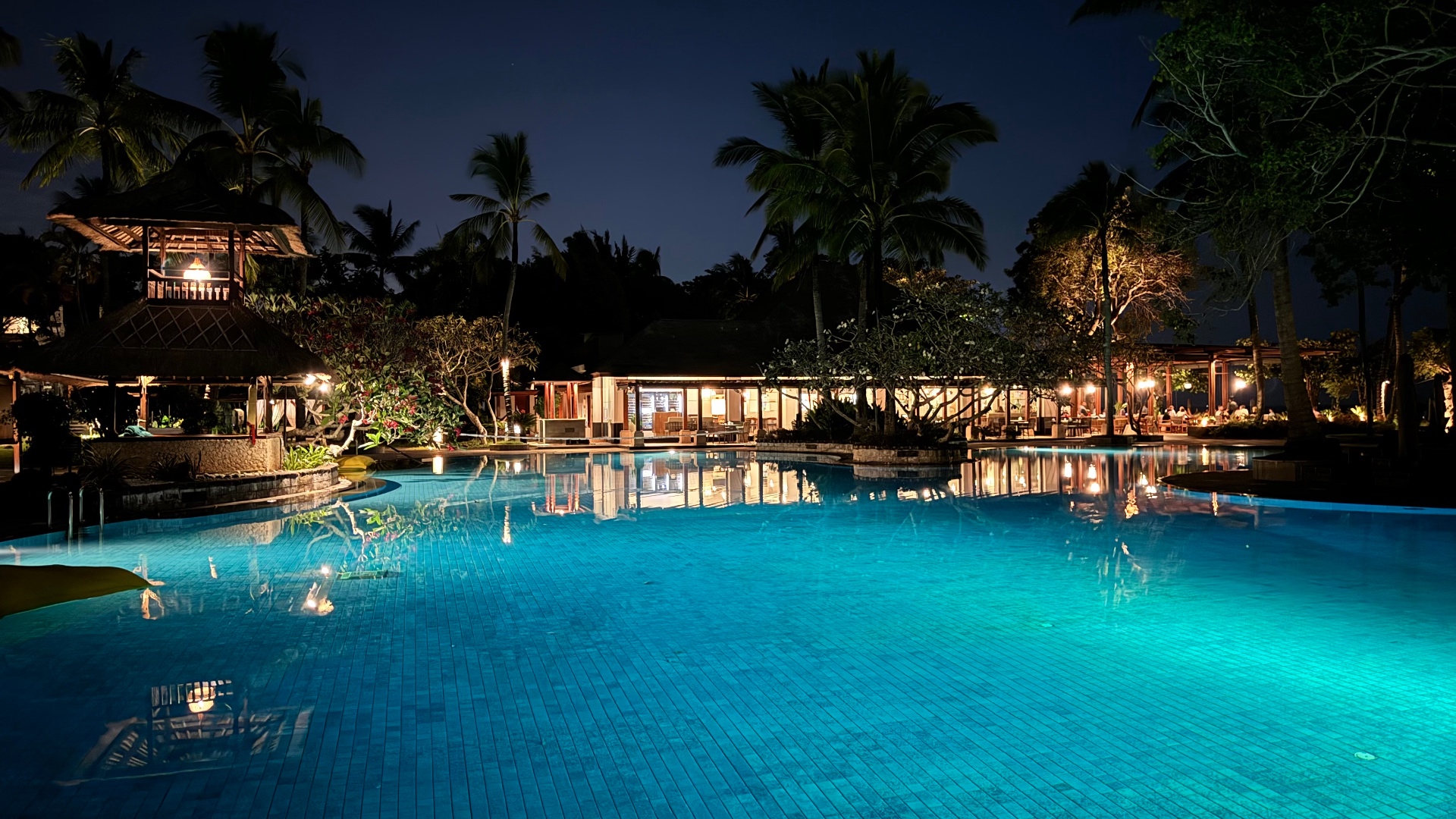 Large turquoise resort pool surrounded by palms and open-air pavilions at night