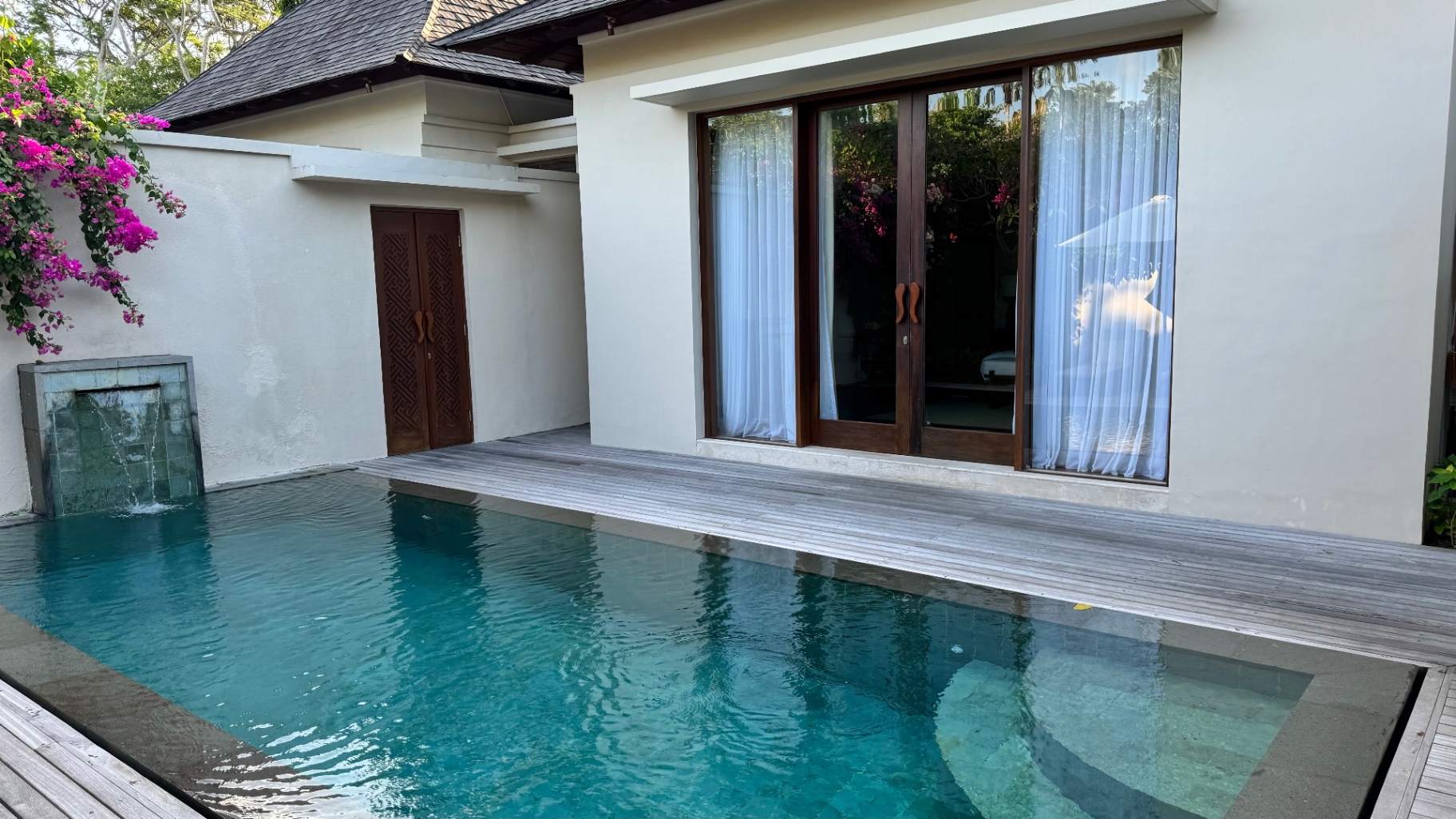 Private villa pool edged by a timber deck, with bougainvillea and a carved door