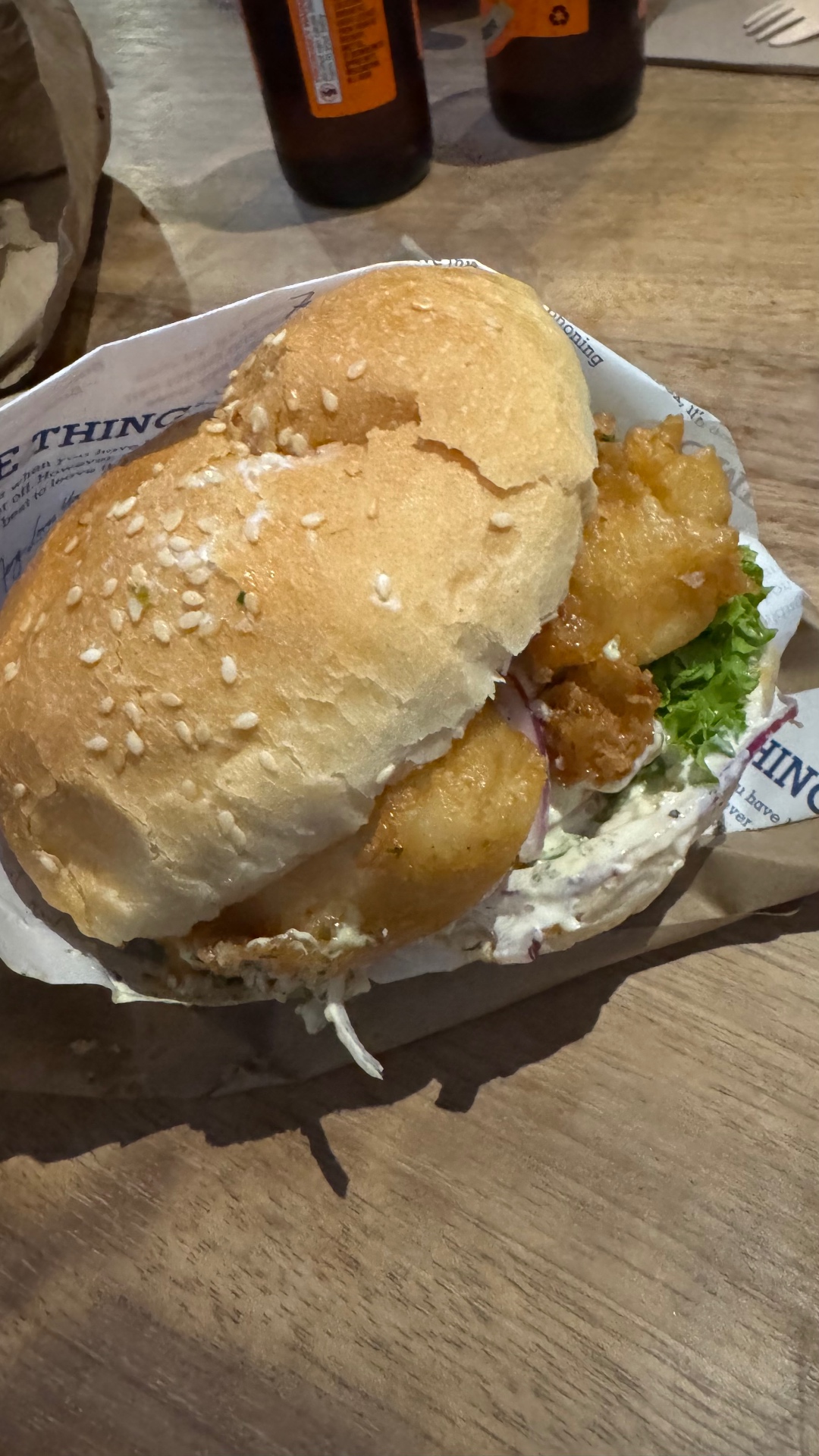 A close-up of a fried chicken burger on a wooden table, with drinks blurred in the background