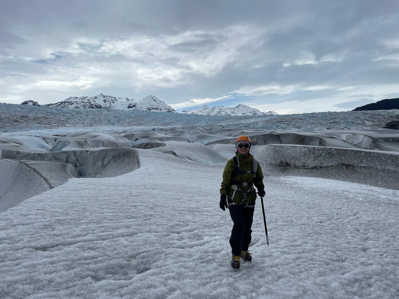 A woman in an olive jacket and orange helmet stands on a vast textured glacier holding a trekking pole, with snow-capped peaks in the distance