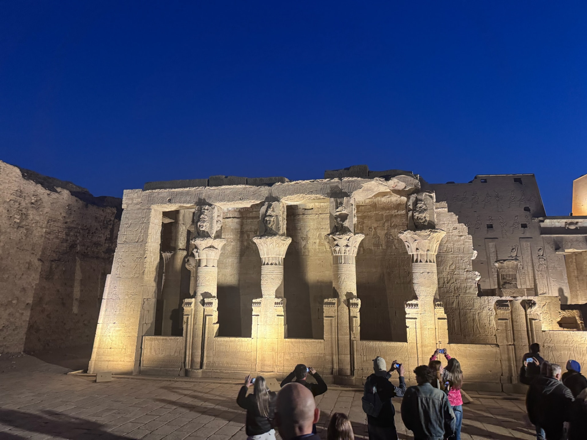 The carved colonnade of Edfu lit gold against a deep blue night sky while visitors photograph it