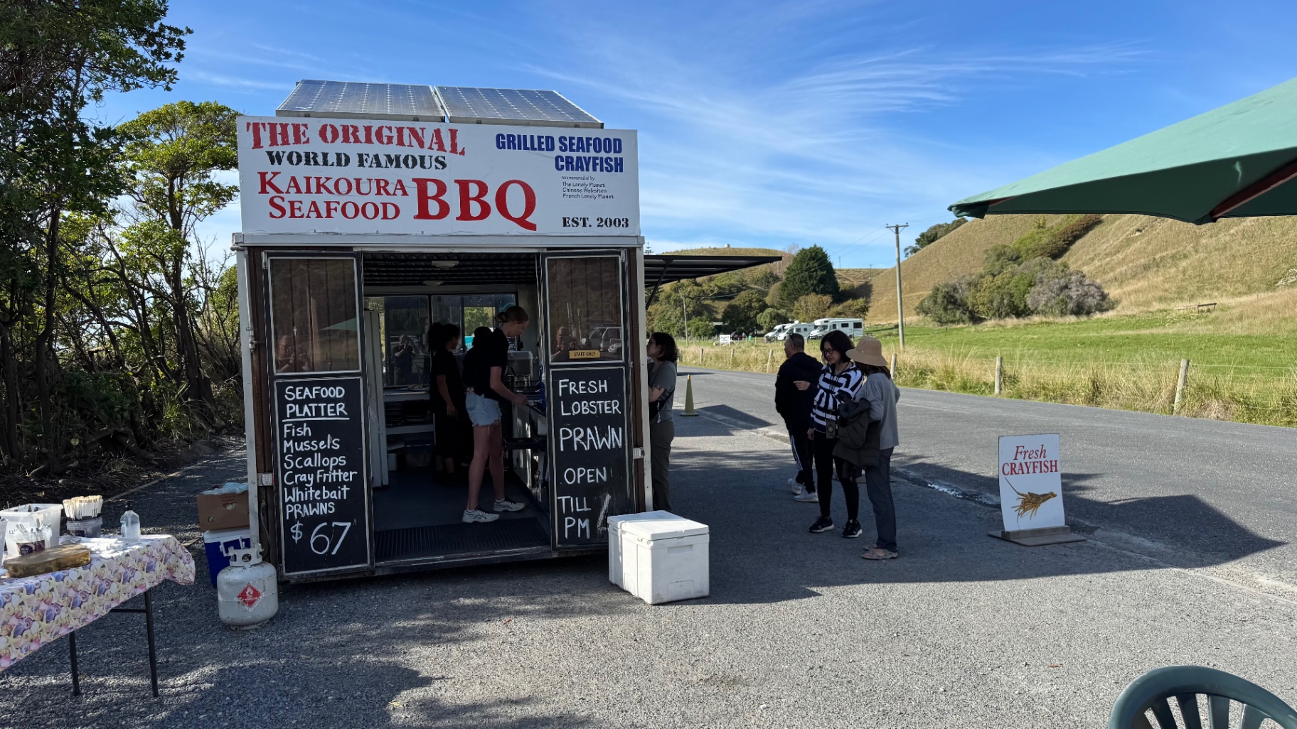 The Original World Famous Kaikoura Seafood BBQ caravan by the roadside, with customers waiting outside under a blue sky