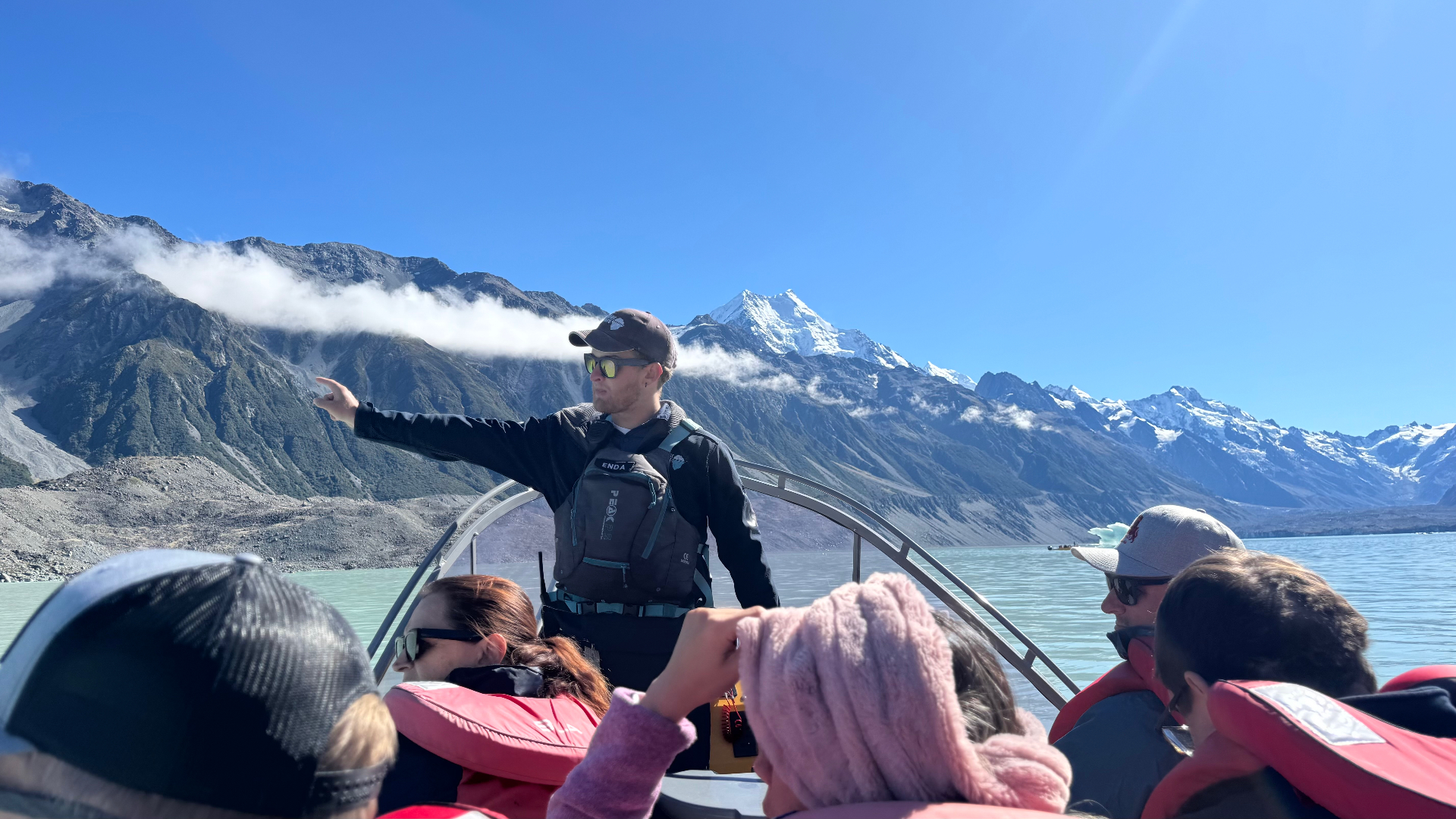 A tour guide standing at the front of a boat pointing towards the mountains, with snow-capped Aoraki / Mount Cook behind him