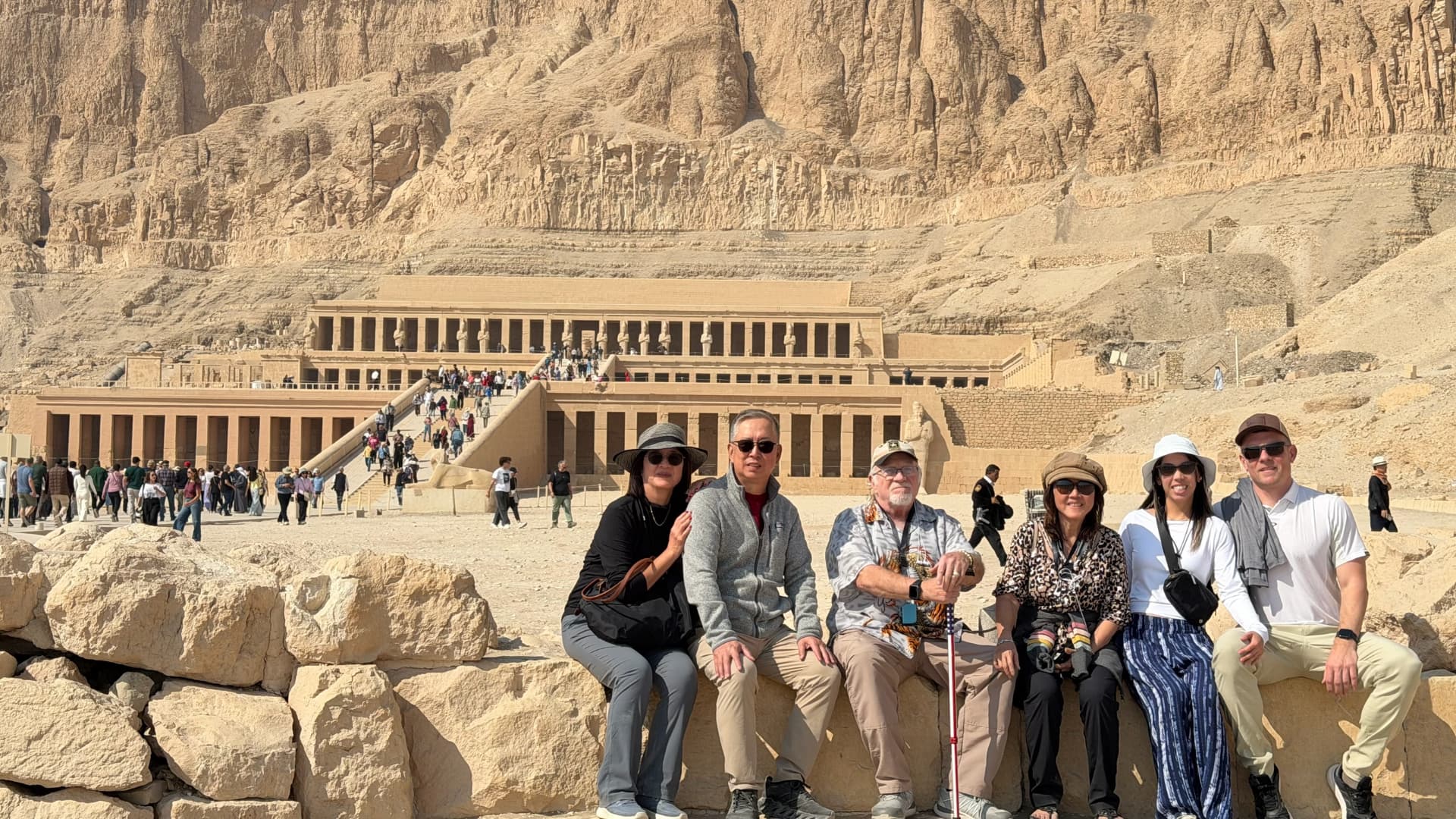Our group sitting on a stone wall with the terraced temple of Hatshepsut and its cliff behind