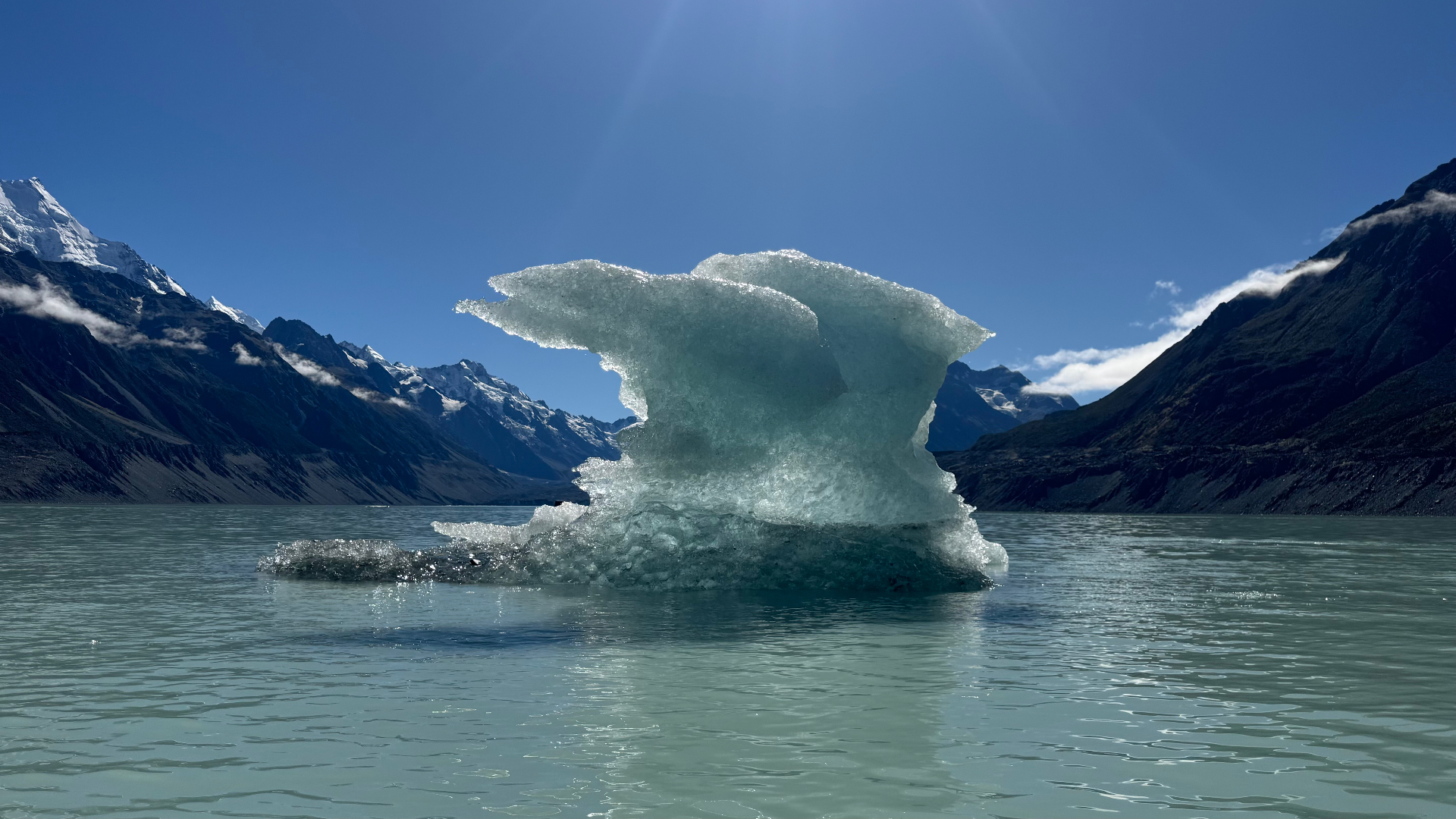 A sculpted iceberg backlit by the sun in the middle of the glacial lake, framed by dark mountains