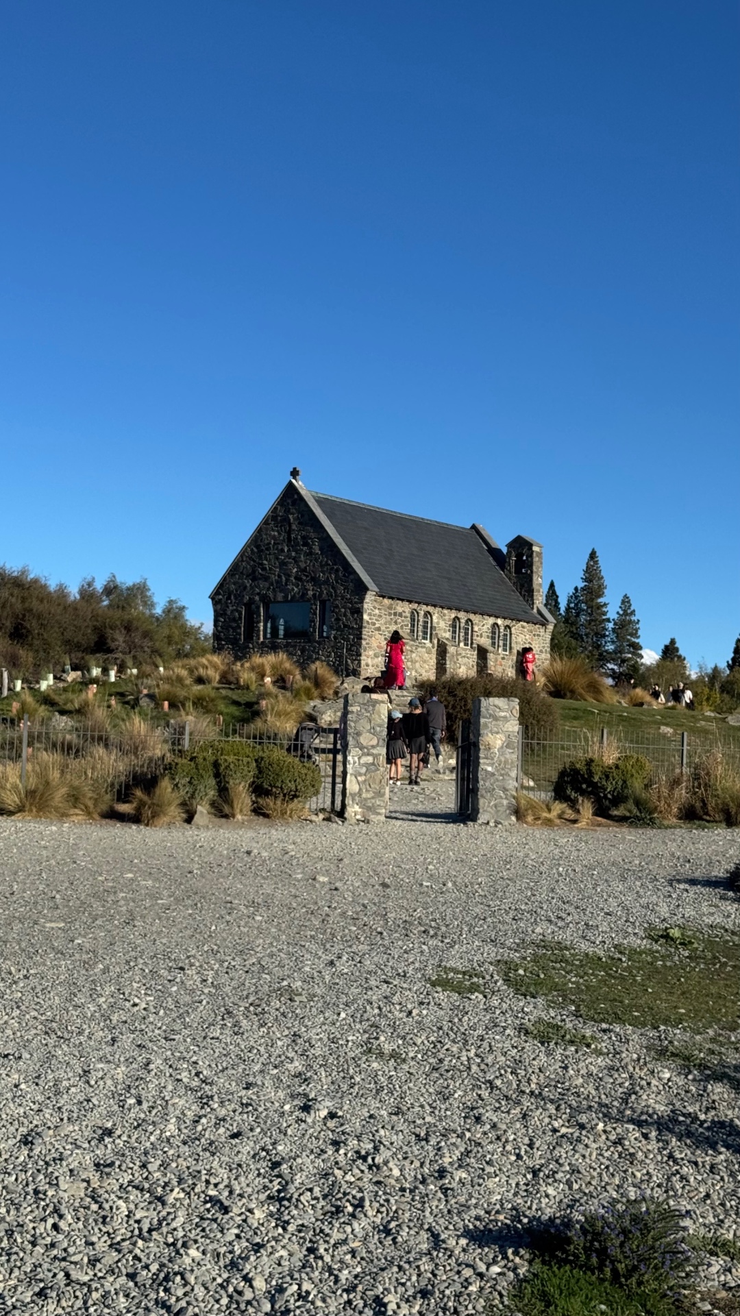 The stone Church of the Good Shepherd on a tussock hillside with visitors walking up through the gate