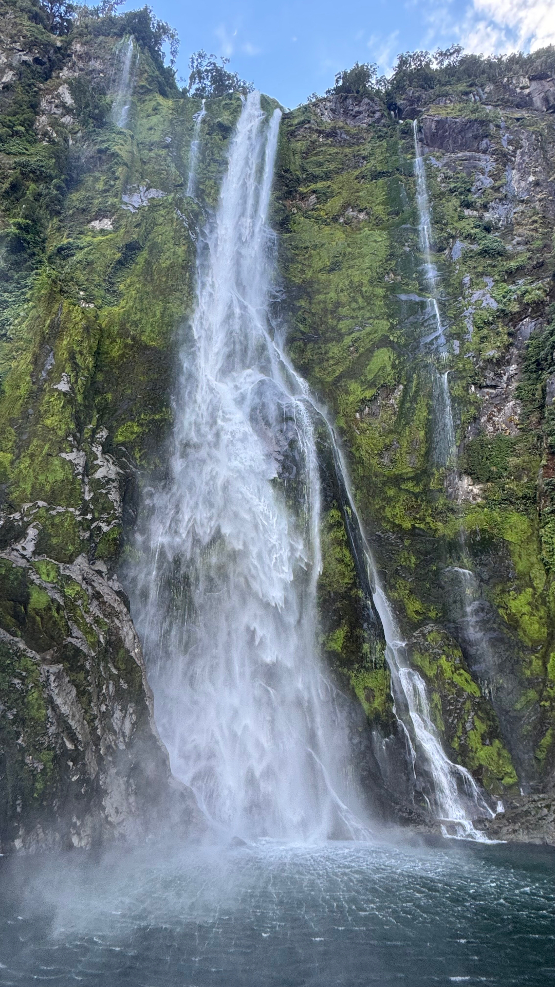 A vertical view of a tall waterfall plunging down a moss-covered cliff face into deep water
