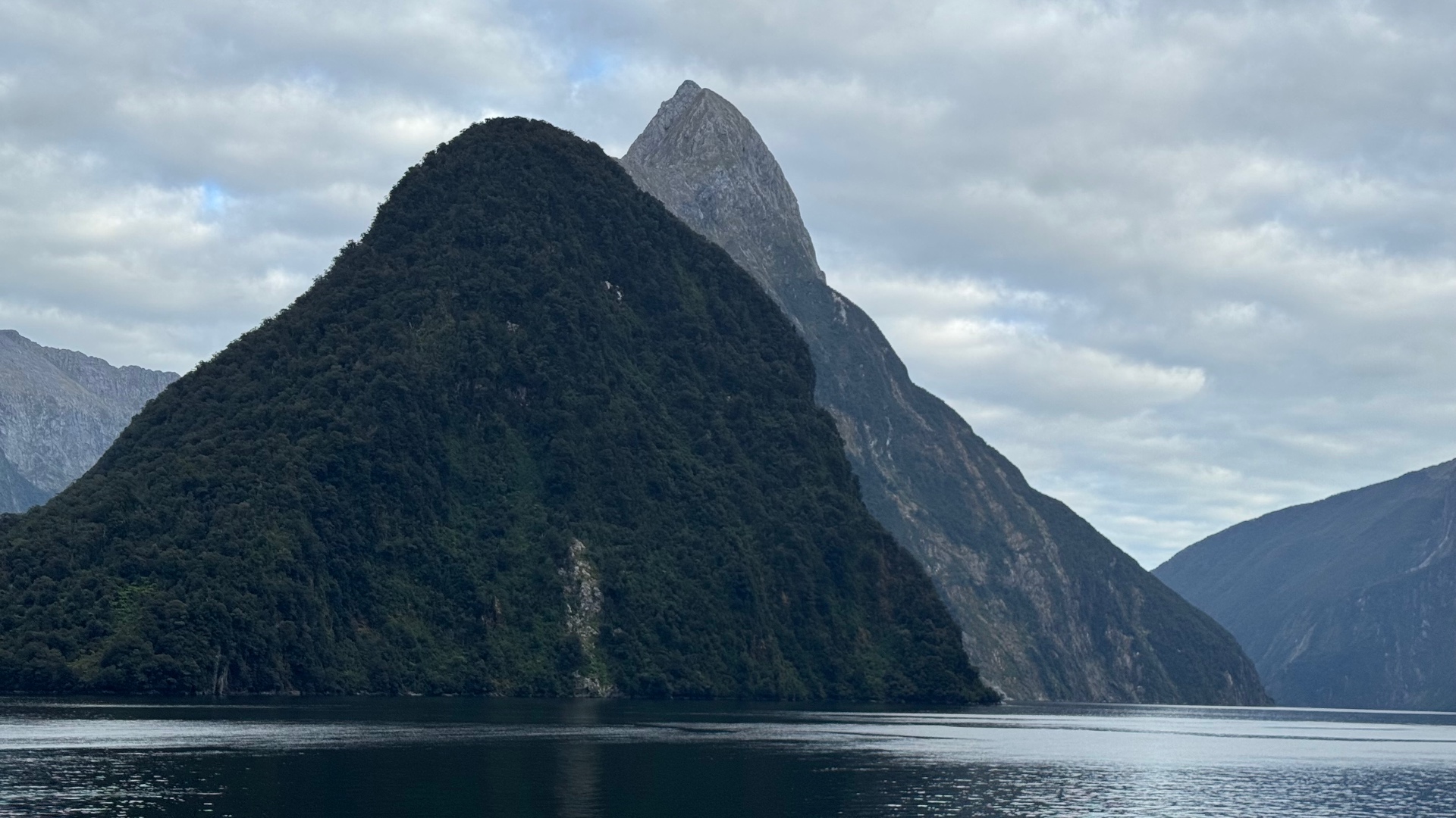 A layered view of Mitre Peak rising behind a lower, densely forested mountain at the water's edge